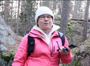 Anne-Maria Paakkola vaalea pipo ja pinkki takki päällään metsässä, kädessään älypuhelin.