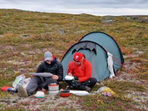 Ida tutkii karttaa ja Ilona kirjoittaa muistiinpanoja istuen teltan edessä.